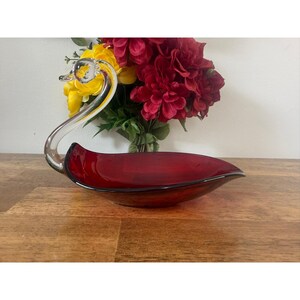 Puede incluir: Un plato en forma de cisne de cristal rojo rubí con cuello y cabeza de cristal transparente. El plato está sobre una superficie de madera, con un ramo de flores rojas y amarillas en el fondo. Es un objeto decorativo.