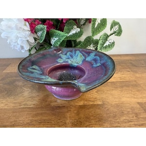 May include: A ceramic flower frog with a wide, shallow dish in shades of purple, teal, and green. The dish has a central opening with a floral pin holder. The frog sits on a short, rounded base. Decorative floral arrangement.
