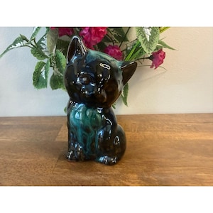 May include: A ceramic cat ornament with a glossy finish. The cat is mainly black with teal and brown accents. It is sitting upright with its ears perked up. The ornament is on a wooden surface, with flowers in the background.