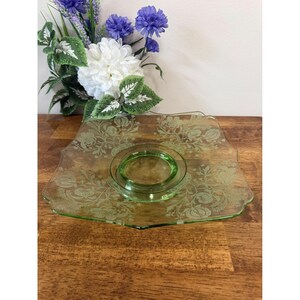 May include: A green glass serving dish with a floral design. The square dish has a scalloped edge and a circular recess in the centre. The dish is on a wooden surface, with flowers in the background.