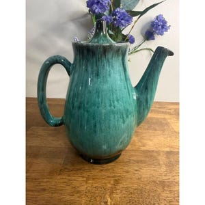 May include: A ceramic teapot featuring a glossy glaze with a blend of deep teal, turquoise, and dark brown vertical drip patterns. The teapot has a curved handle, a pointed spout, and a matching lid with a tapered finial top.