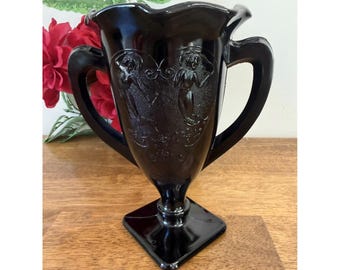Peut inclure: Vase en verre noir avec deux poignées et une base carrée. Le vase présente des figures en relief et un bord festonné. Le vase est présenté sur une surface en bois avec des accents floraux rouges et verts en arrière-plan.