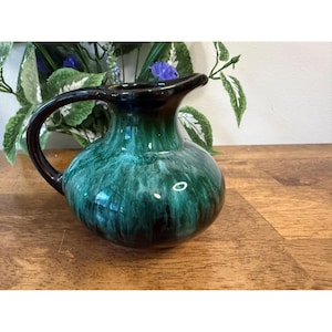 May include: A ceramic jug with a dark handle and rim. The jug has a glossy finish with shades of green and black. The jug is sitting on a wooden surface.