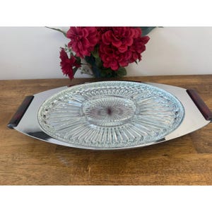 Può includere: Un vassoio ovale da portata con un inserto in vetro trasparente e una cornice in metallo color argento. L'inserto in vetro ha un motivo a stella. Il vassoio ha manici marroni scuri su ciascuna estremità. Fiori rossi sullo sfondo.