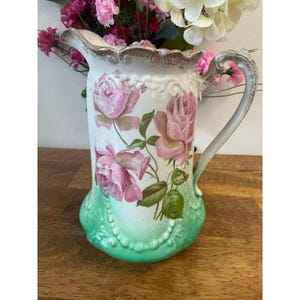 Peut inclure: Pichet en céramique blanche orné de motifs floraux de roses roses et d'accents verts. Le pichet a un bord festonné avec une garniture dorée et une poignée incurvée. La base du pichet est de couleur vert clair avec un motif de perles.