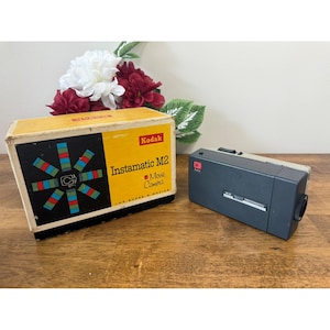 May include: A vintage Kodak Instamatic M2 Movie Camera and its original box. The box is yellow with black and colorful geometric designs. The camera is dark gray with a silver top and black lens.