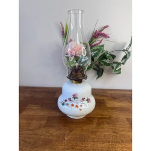 Puede incluir: Una lámpara de aceite vintage con una base de cerámica blanca adornada con motivos florales. La chimenea de vidrio transparente alberga una flor rosa. La lámpara tiene un accesorio de metal color latón y está sobre una superficie de madera.