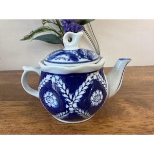 Könnte beinhalten: Eine weiße Keramik-Teekanne mit tiefblauer Glasur und einem weißen Blumen- und Blattmuster. Die Teekanne verfügt über einen gewellten Rand, einen geschwungenen Griff, einen kurzen Ausguss und einen passenden Deckel mit einem kleinen Schlaufengriff.
