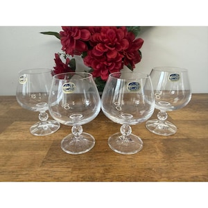 May include: Four clear crystal brandy snifters with round bowls and short stems are arranged on a wooden surface. Each glass has a small, round base and a decorative stem. A bouquet of red flowers is in the background.