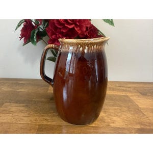 May include: A brown ceramic pitcher with a handle, featuring a glossy finish and a lighter brown rim. The pitcher is displayed on a wooden surface, with red flowers in the background.