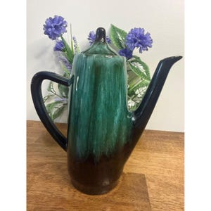 May include: A tall, ceramic coffee pot with a dark green and brown glaze. The handle and spout are black. The pot is displayed on a wooden surface with purple flowers in the background.