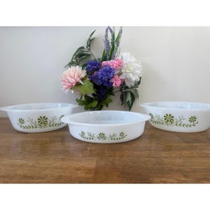 May include: Three white oval casserole dishes with green floral patterns. The dishes are on a wooden surface, with a bouquet of flowers in the background. Each dish has handles.