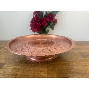 May include: A copper-coloured, round cake stand with a scalloped edge and an etched pattern. The stand has a pedestal base. A bouquet of red flowers is in the background. The stand is displayed on a wooden surface.