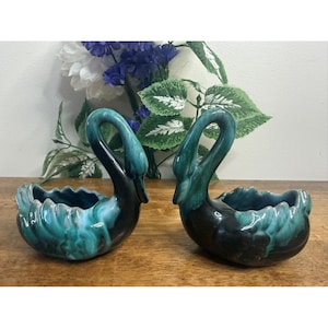 Puede incluir: Dos maceteros de cerámica con forma de cisne, esmaltados en verde azulado y negro. El cuello del cisne se curva con elegancia, y el cuenco abierto tiene un borde festoneado. Flores decorativas visibles en el fondo. Un artículo de decoración vintage.