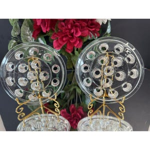 May include: Two clear glass plates with circular designs and multiple holes, presented on ornate gold-coloured stands. The plates are set against a backdrop of red flowers and greenery, creating a decorative display.
