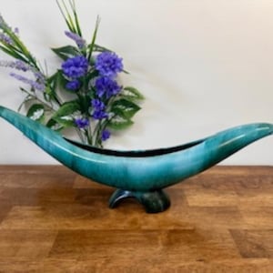 May include: A teal ceramic centerpiece bowl with a curved, elongated design. The bowl is supported by a three-footed base and filled with purple flowers and greenery. The bowl's interior is black.