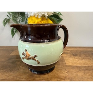 Può includere: Brocca in ceramica marrone e verde con manico e beccuccio. La brocca presenta una fascia verde chiaro con un rilievo in rilievo di una persona a cavallo. La parte superiore e inferiore sono marroni.