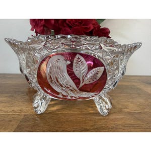 May include: A clear, footed crystal bowl with a ruby red oval centre featuring an etched bird and leaf design. The bowl has a diamond-cut pattern and is filled with red flowers.