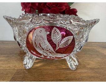 May include: A clear, footed crystal bowl with a ruby red oval center featuring an etched bird and leaf design. The bowl has a diamond-cut pattern and is filled with red flowers.