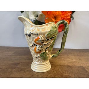 May include: Cream-colored ceramic pitcher with a textured surface and a flared spout. The pitcher features a handle designed to resemble a tree branch and is decorated with acorn and leaf motifs in orange, green, and black.