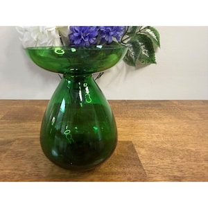 Peut inclure: Un vase en verre vert vif, avec un large bol peu profond sur le dessus et une base en forme de goutte d'eau. Le vase contient des fleurs blanches et violettes et repose sur une surface en bois. Le verre reflète la lumière.