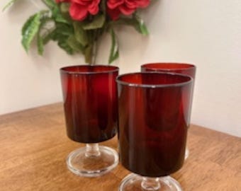 May include: Three red glass goblets with clear glass stems and bases. The goblets are on a wooden surface.
