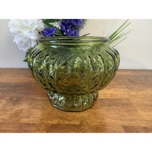 May include: A green glass vase with a textured, diamond-like pattern. The vase has a wide, rounded bowl shape and a pedestal base. Artificial white and purple flowers are visible in the background. The vase is sitting on a wooden surface.