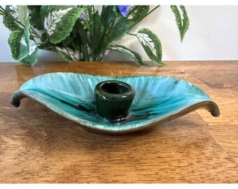 Puede incluir: Un plato de cerámica en forma de hoja, de color turquesa y verde, con un portavelas cilíndrico verde oscuro en el centro. El plato tiene un acabado brillante y un borde marrón. Arreglo floral decorativo en el fondo.