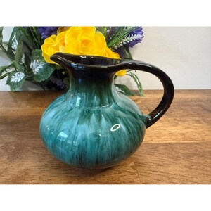 Op de afbeelding: Een keramische kan met een afgeronde body en een donkere handgreep en tuit. De kan heeft een glanzende afwerking met een teal en donkergroen kleurenschema. De kan staat op een houten oppervlak.