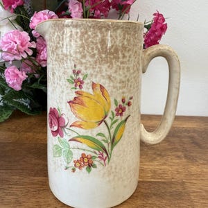 Puede incluir: Jarra de cerámica vintage con acabado moteado beige y asa grande. La jarra está decorada con un diseño floral con un tulipán amarillo, una rosa roja y otras flores coloridas. La jarra probablemente se utiliza para servir bebidas.