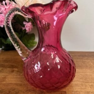 May include: A ruby-red glass jug with a textured, diamond-patterned body and a ruffled rim. The clear handle is arched. The jug is displayed on a wooden surface, with flowers in the background.