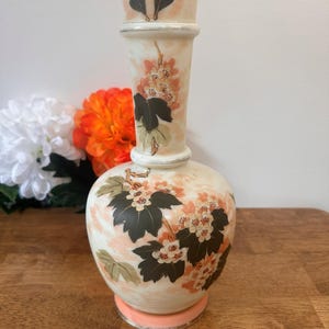 May include: A decorative ceramic vase with a bulbous base and a tall, cylindrical neck. The vase has a cream-coloured background and features floral designs in shades of peach, brown, and green. The base is a light peach colour.
