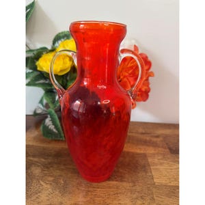 May include: A striking red glass vase with two arched handles. The vase has a textured finish and a broad opening. The background includes yellow and orange flowers, adding colour. This decorative vase is suitable for floral arrangements and home decor.