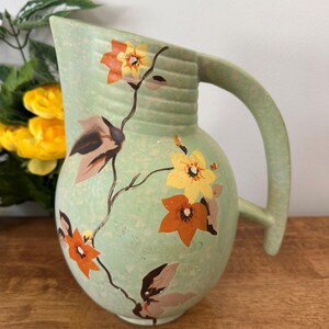 Könnte beinhalten: Eine hellgrüne Vintage-Kanne mit großem Griff und Ausguss. Die Kanne ist mit einem Blumendesign mit orangefarbenen, gelben und braunen Blumen und Blättern verziert. Die Kanne steht auf einer Holzoberfläche.