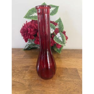 May include: A slender, ruby red glass vase with vertical ribbed detailing. The vase has a narrow neck and a rounded base. The background features red flowers and green leaves, suggesting a decorative floral arrangement.
