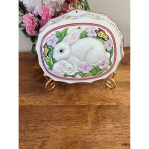 May include: White ceramic wall plaque with a raised relief design of two rabbits surrounded by flowers and butterflies. The plaque has a pink border and is displayed on a gold-coloured stand. A floral arrangement is in the background.