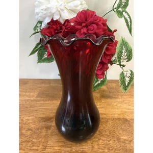 Peut inclure: Un vase en verre rouge foncé avec un bord festonné. Le vase a une base bulbeuse et un col fin. Le vase est rempli de fleurs rouges et blanches et de feuillage vert. Le vase est posé sur une surface en bois.