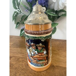 Puede incluir: Una jarra de cerveza vintage con tapa y asa plateadas. El cuerpo de cerámica presenta una escena detallada y colorida de personas en una mesa. La jarra tiene una combinación de colores marrón, naranja y azul.
