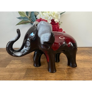 Puede incluir: Una hucha con forma de elefante de cerámica, en tonos marrón y rojo con detalles blancos. El elefante tiene una larga trompa, orejas grandes y una ranura para monedas. Un artículo decorativo.