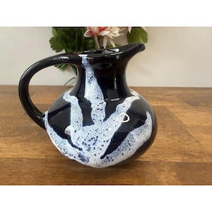 May include: A dark blue ceramic jug with a white, abstract design. The jug has a curved handle and a spout for pouring. The white design appears to be splattered across the surface of the jug. The jug is sitting on a wooden surface.