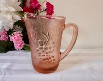 May include: Pink glass pitcher with a textured surface and a handle. The pitcher features a grape design on the front. The pitcher is displayed on a white surface with flowers in the background.