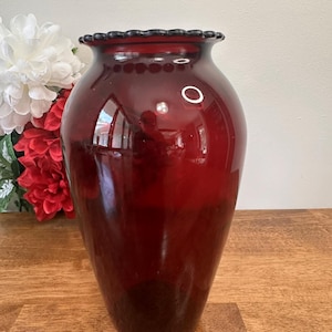 May include: A deep red glass vase with a decorative, ruffled rim. The vase has a rounded body and stands on a wooden surface. White and red flowers are in the background. A decorative item.