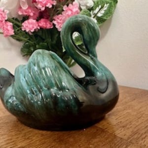 Puede incluir: Un macetero de cerámica esmaltada en forma de cisne, en tonos verdes y negros. El cuello del cisne está curvado y su cabeza está inclinada hacia abajo. El macetero es perfecto para exhibir plantas o flores pequeñas.