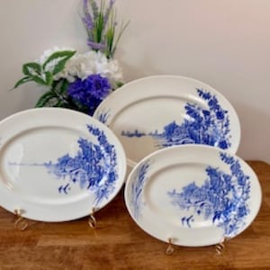 May include: Three white oval serving platters with a blue landscape design. The platters are displayed on gold-coloured stands. The largest platter is approximately 40 cm long, the medium platter is 30 cm long, and the smallest is 23 cm long. Floral arrangement in the background.