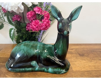 May include: A ceramic deer figurine in a reclining pose. The deer has a glossy, dark green and black glaze. The ears are upright, and the body is smooth. The figurine is displayed on a wooden surface.