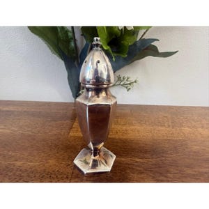 Pode incluir: Um saleiro de metal prateado com base hexagonal, corpo cónico com painéis verticais planos e tampa superior arredondada e perfurada. O objeto tem uma superfície polida e refletora, posicionado sobre uma superfície de madeira.