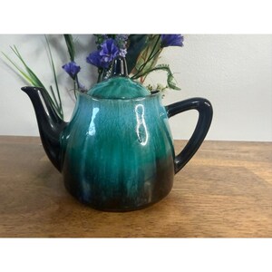 Può includere: Una teiera in ceramica con una smaltatura lucida che sfuma da una base nera profonda a un centro vibrante color turchese e verde acqua. La teiera include un coperchio abbinato con un piccolo pomello, un beccuccio curvo e un manico robusto.