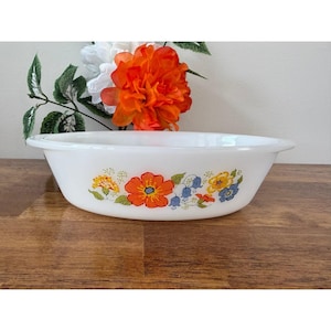 Puede incluir: Fuente ovalada blanca con un diseño floral con flores naranjas, azules y amarillas. El plato tiene una forma simple y redondeada y está sobre una superficie de madera. Las flores están dispuestas en un patrón repetitivo.