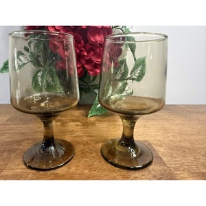 Può includere: Due calici da vino color ambra con una coppa arrotondata e uno stelo. I bicchieri sono in vetro trasparente e poggiano su una superficie di legno. Un bouquet di fiori rossi è sullo sfondo.