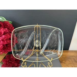 Puede incluir: Una fuente de vidrio transparente rectangular con diseños florales grabados, exhibida en un soporte de metal dorado. La fuente tiene dos secciones curvas y un motivo floral central. El soporte tiene una base decorativa.